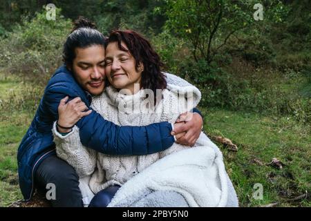 Positive Mutter und Sohn in warmen Klamotten, die auf Gras sitzen und sich gegenseitig in grünen Wäldern umarmen Stockfoto
