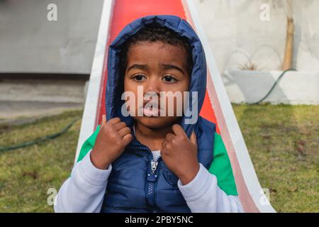 Fokussierter kleiner afroamerikanischer Junge in warmer Weste mit Kapuze, die auf roten Kindern sitzt, rutscht auf dem Spielplatz und schaut weg Stockfoto