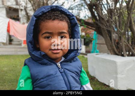 Fokussierter kleiner afroamerikanischer Junge in warmer Weste mit Kapuze auf dem Spielplatz und mit Blick weg Stockfoto