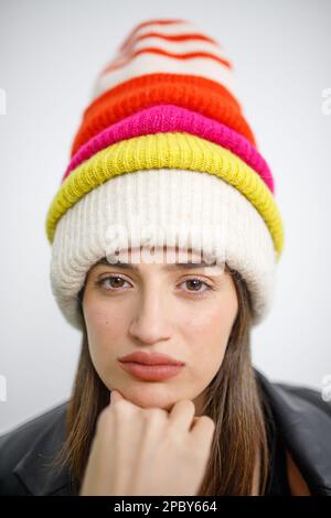 Porträt einer jungen Frau in warmer Kleidung und farbenfrohen Strickmützen, die vor weißem Hintergrund in die Kamera schaut Stockfoto