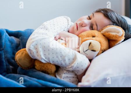 Süßes Mädchen im Schlafanzug, das auf einem weichen weißen Kissen schläft und ein weiches Spielzeug umarmt Stockfoto