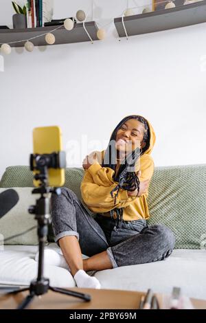 Fröhliche junge afroamerikanische Bloggerin in lässigem, bequemem Outfit mit Zöpfen, die sich selbst umschließen und beim Aufnehmen von Podcas auf den Bildschirm schauen Stockfoto