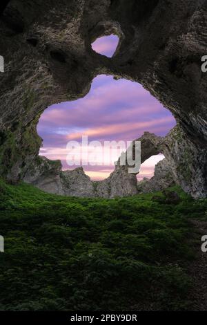 Arco de Portupekoleze Höhle mit Doppelloch in Felsformationen mit grünem Gras in malerischer Landschaft vor farbenfrohem Sonnenuntergang in Spanien Stockfoto