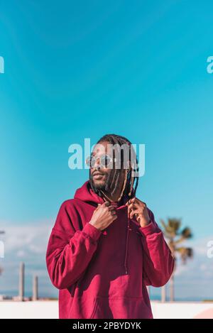 Seriöser afroamerikanischer Mann mit rotem Hoodie und Sonnenbrille mit Dreadlocks, der vor dem blauen Himmel auf die Kamera schaut Stockfoto