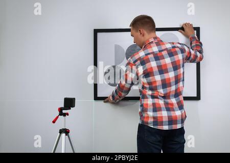 Mann, der eine Kreuzlinienlaser-Wasserwaage zum Aufhängen von Malerarbeiten an eine helle Wand verwendet, Rückansicht Stockfoto