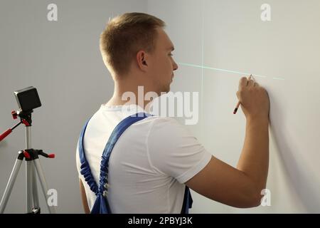 Arbeiter, der Kreuzlinienlaser-Nivelliergerät und Bleistift für genaue Messungen an hellen Wänden verwendet Stockfoto