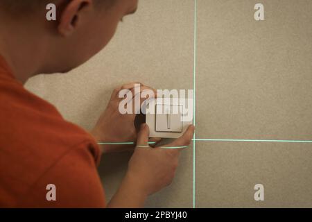 Mann mit Kreuzlinien-Laser-Nivelliergerät zur Installation eines Lichtschalters in Gipskartonplatten Stockfoto