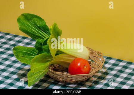 Frischer grüner Bok Choy Kohl und rote reife, saftige Tomate auf karierte Tischdecke vor gelbem Hintergrund gelegt Stockfoto