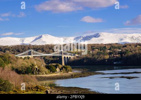 Malerischer Blick auf die Menai-Hängebrücke, die die Menai-Straße überquert, mit Schnee auf den Bergen im Winter. Menai Bridge (Porthaethwy), Isle of Anglesey, Wales, Vereinigtes Königreich Stockfoto