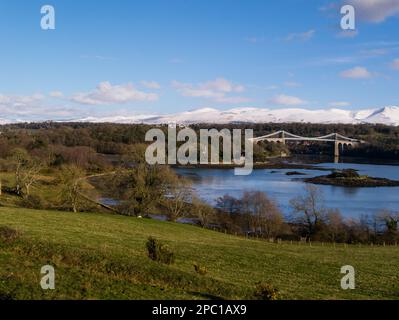 Blick über die Menai-Straße zur Menai-Brücke und Kircheninsel mit St. Tysilio's-Kirche und schneebedeckten Carnedau Mountains im Hintergrund auf hellen s Stockfoto