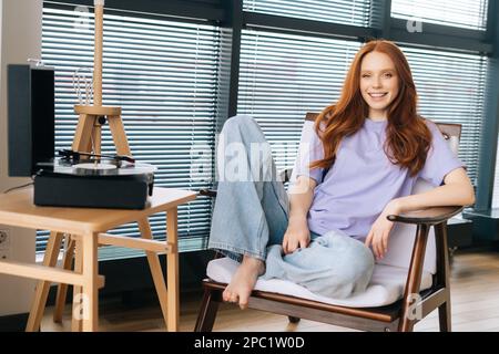 Porträt einer fröhlichen jungen Frau, die Musik auf einem alten Grammophon-Schallplattenspieler im hellen Wohnzimmer hört und auf einem Stuhl im Hintergrund sitzt Stockfoto