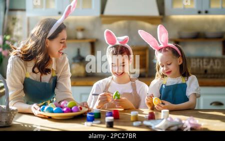 Fröhliche Mutter und ihre Töchter tragen Schürzen mit bemalten, farbenfrohen Eiern, während sie sie in der gemütlichen Küche zu Hause mit Färbemitteln dekorieren. Osterfahrzeuge Stockfoto