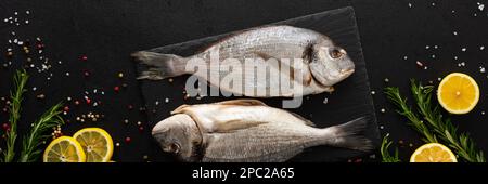 Dorada-Fisch mit Zitronenscheiben und Rosmarin-Zweigen auf einem Schieferbrett mit Gewürzen auf einem schwarzen Banner, Draufsicht, Kopierbereich Stockfoto