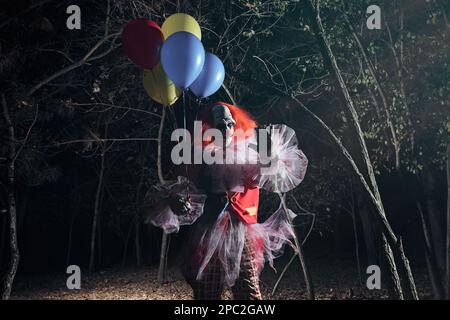 Furchteinflößender Clown mit Luftballons draußen in der Nacht. Halloween-Party-Kostüm Stockfoto