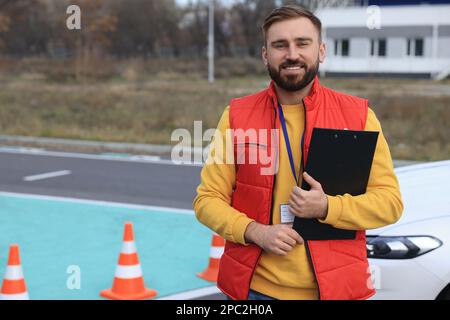 Kursleiter mit Klemmbrett in der Nähe des Fahrzeugs auf der Teststrecke. Fahrschule Stockfoto