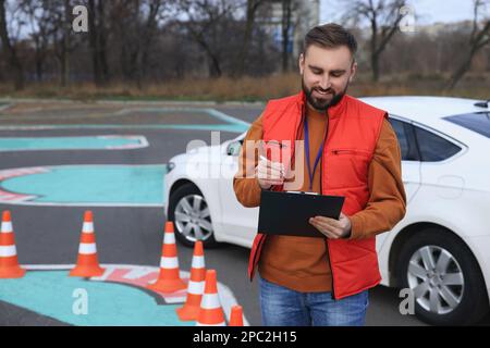 Kursleiter mit Klemmbrett in der Nähe des Fahrzeugs auf der Teststrecke. Fahrschule Stockfoto