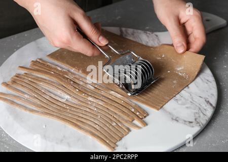 Frau, die Soba (Buchweizennudeln) mit Schneidevorrichtung am grauen Tisch macht, Nahaufnahme Stockfoto