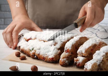 Eine Frau schneidet traditionelle Weihnachtsstollen mit Puderzucker auf einem weißen Holztisch, Nahaufnahme Stockfoto