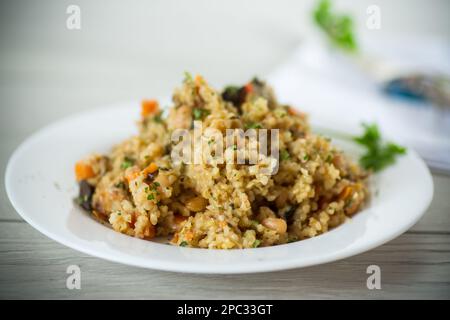 Gekochter Bulgur mit Gemüse, Karotten und getrockneten Pilzen auf einem Teller auf einem Holztisch. Stockfoto