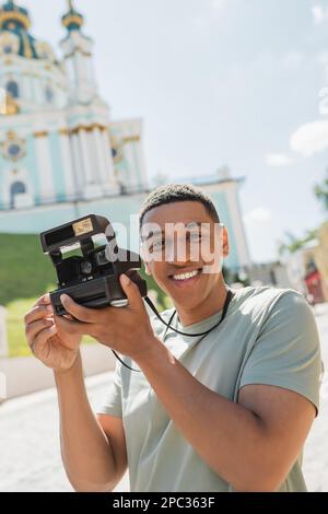 Sorgenfreier afroamerikanischer Tourist mit einer antiken Kamera, die in der Nähe der verschwommenen St. Andrews Kirche in Kiew lächelt, Stockbild Stockfoto