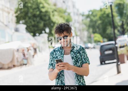 Überglücklich Mann mit Sonnenbrille läuft und benutzt Smartphone auf der Straße, Stock Image Stockfoto