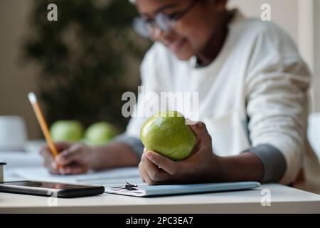 Konzentrieren Sie sich auf die Hand eines fleißigen Schuljungen, der frischen grünen Apfel hält, während er zu Hause am Schreibtisch sitzt und Hausaufgaben erledigt Stockfoto