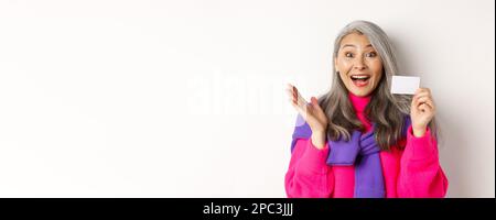 Einkaufskonzept. Glücklich asiatische alte aldy suchen beeindruckt und zeigt Kunststoff-Kreditkarte ihrer Bank, über weißem Hintergrund stehen Stockfoto