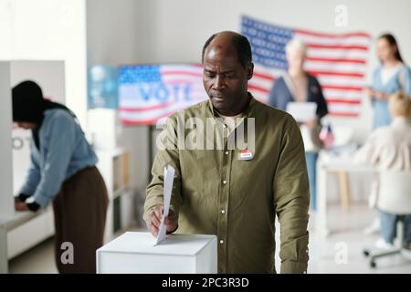 Reifer afroamerikanischer Mann, der an Präsidentschaftswahlen teilnimmt, während er an der Wahlurne steht und ein Dokument mit seiner Wahl dort ablegt Stockfoto