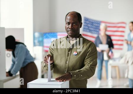 Ein reifer Schwarzer sieht vor die Kamera, während er bei Präsidentschaftswahlen Papier mit seinem gewählten Kandidaten in die Wahlurne legt Stockfoto