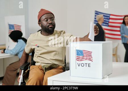 Afroamerikanischer junger Mann mit Behinderung, der im Rollstuhl sitzt und Stimmzettel in den Karton legt, wobei die US-Flagge auf dem Schreibtisch steht Stockfoto