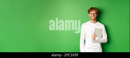 Hübscher junger Mann mit roten Haaren, Brille und Langarm-T-Shirt, Laptop auf grünem Hintergrund haltend Stockfoto