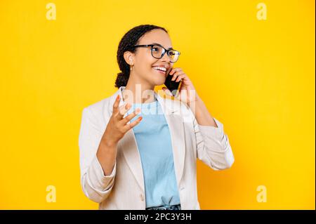 Positive Begeisterung erfolgreiche vielbeschäftigte brasilianerin oder hispanische Frau, die mit einem Freund oder Kollegen telefoniert, wegschaut, Gesten mit der Hand macht, lächelt, auf isoliertem gelbem Hintergrund steht Stockfoto