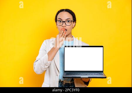 Überraschend erfolgreiche braune Geschäftsfrau mit gemischten Rassen, halten Sie Ihren Laptop mit einem leeren weißen Modell-Kopierbildschirm, stehen Sie auf einem isolierten gelben Hintergrund, und schauen Sie in Erstaunen auf die Kamera Stockfoto