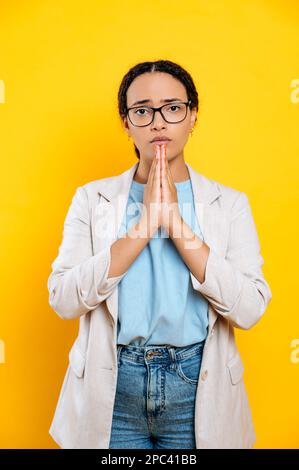 Ich bitte um etwas, bete. Vertikales Foto einer süßen gemischten Rasse, Arbeiterin, hält Hände in einem gefalteten Gebetsschild, schaut in die Kamera, steht auf isoliertem gelben Hintergrund Stockfoto