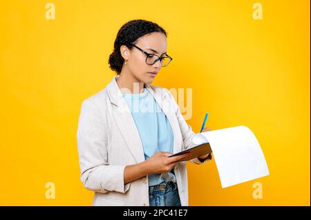 Fokussierte, erfolgreiche braune Frau mit gemischter Rasse, trägt stilvolle Freizeitkleidung, HR Manager, Immobilienmakler, hält und unterschreibt Dokumente, konzentriert auf sie, steht auf isoliertem gelbem Hintergrund Stockfoto