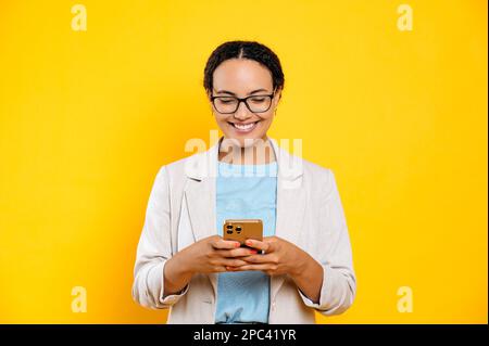 Positiv erfolgreiche vielbeschäftigte hispanische oder brasilianische Frau, die ihr Smartphone nutzt, mit einem Freund oder Kollegen in sozialen Medien SMS schreibt, auf den Bildschirm schaut, lächelt, auf isoliertem gelbem Hintergrund steht Stockfoto