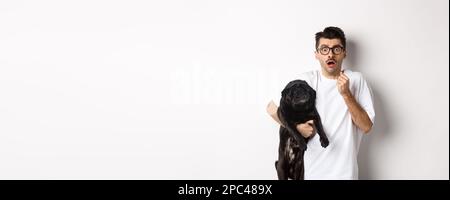 Erschrockener Hipster umarmt seine niedlichen schwarzen Welpen und starrte auf Kamera erschrocken. Hundebesitzer schaut schockiert, hält Mops, steht auf weißem Hintergrund Stockfoto