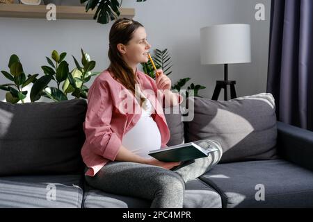 Traumhaft glückliche, lächelnde junge schwangere Frau, die auf einem bequemen Sofa sitzt und zu Hause in ein Tagebuch oder ein Notizbuch schreibt. Schöne zukünftige Mutter, die Shopp macht Stockfoto