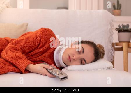 Junge Frau in orangefarbenem Pullover mit orthopädischem Kragen, die die Fernbedienung des Fernsehers hält, während sie zu Hause auf einem bequemen Sofa im Wohnzimmer schläft Stockfoto