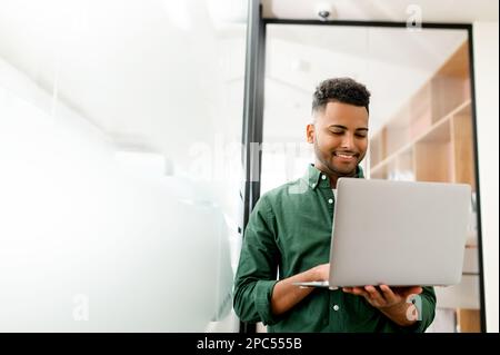 Porträt eines charismatischen, eleganten, jungen indischen Geschäftsmanns, der im modernen Büro steht und ein Laptop benutzt, und männlicher arabischer Freiberufler genießt die Arbeit in einem Gemeinschaftsarbeitsplatz Stockfoto