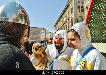 Zwei Clowns in weißen Kostümen sehen einer jungen braunen Eule zu, die auf der Hand seines Besitzers sitzt, gekleidet in einem mittelalterlichen Ritterkostüm mit eisernem Helm. Stockfoto