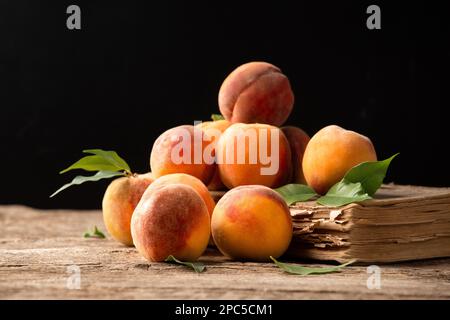 Pfirsiche mit grünen Blättern liegen auf einem Buchhölzer-Hintergrund. Reife Pfirsiche aus der Nähe auf einem alten Holzhintergrund. Frische Pfirsiche auf einem alten Buch. Stockfoto