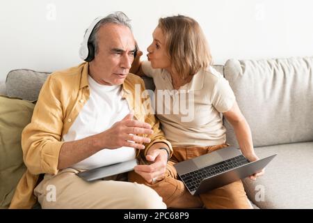 Senior-Mann, der Musik über Kopfhörer hört und sich nicht darum kümmert, dass seine Frau sein Headset abnimmt und mit ihm spricht Stockfoto