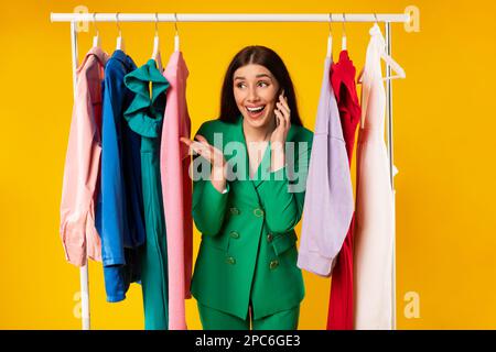 Shoppingnachrichten. Glückliche Shopaholic Frau, die mit dem Handy spricht, zwischen Kleidern in der Nähe des Kleiderständers steht Stockfoto