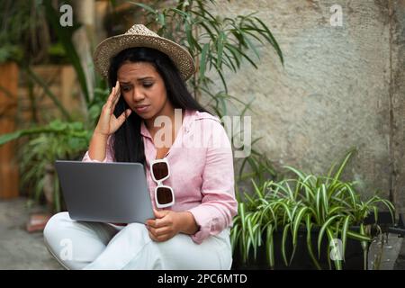 Eine verwirrte, unglückliche, junge schwarze Frau mit Hut sitzt auf dem Boden mit einem Computer und sucht nach einer Route auf der Karte Stockfoto