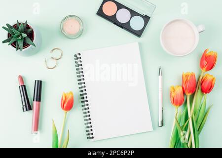 Unbeschrifteter Notizblock, eine Tasse Kaffee, Kosmetika und ein Strauß orangefarbener Tulpen auf hellgrünem Hintergrund, Draufsicht. Frauen- oder Muttertagskonzept. Stockfoto
