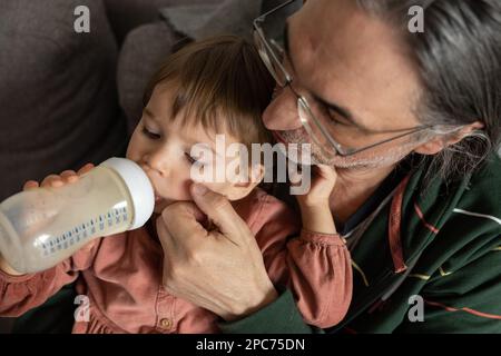 Ein kleines Mädchen trinkt Milch aus einer Flasche, während es auf dem Schoß ihres Großvaters sitzt. Stockfoto