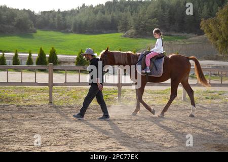 Glückliches kleines Mädchen, das in der Reitschule reitet. Ein Mädchen, das in der Manege reitet, begleitet von einem Reitlehrer. Pferdekonzert. Stockfoto