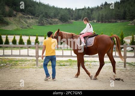 Glückliches kleines Mädchen, das in der Reitschule reitet. Mädchen, die in der Manege reitet, begleitet von einem Reitlehrer. Reitkonzept. Stockfoto