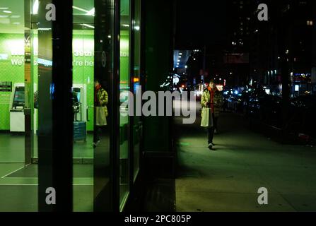 Eine junge Frau, die nachts an der Geldautomatenlobby einer Bank in New York City vorbeigeht Stockfoto